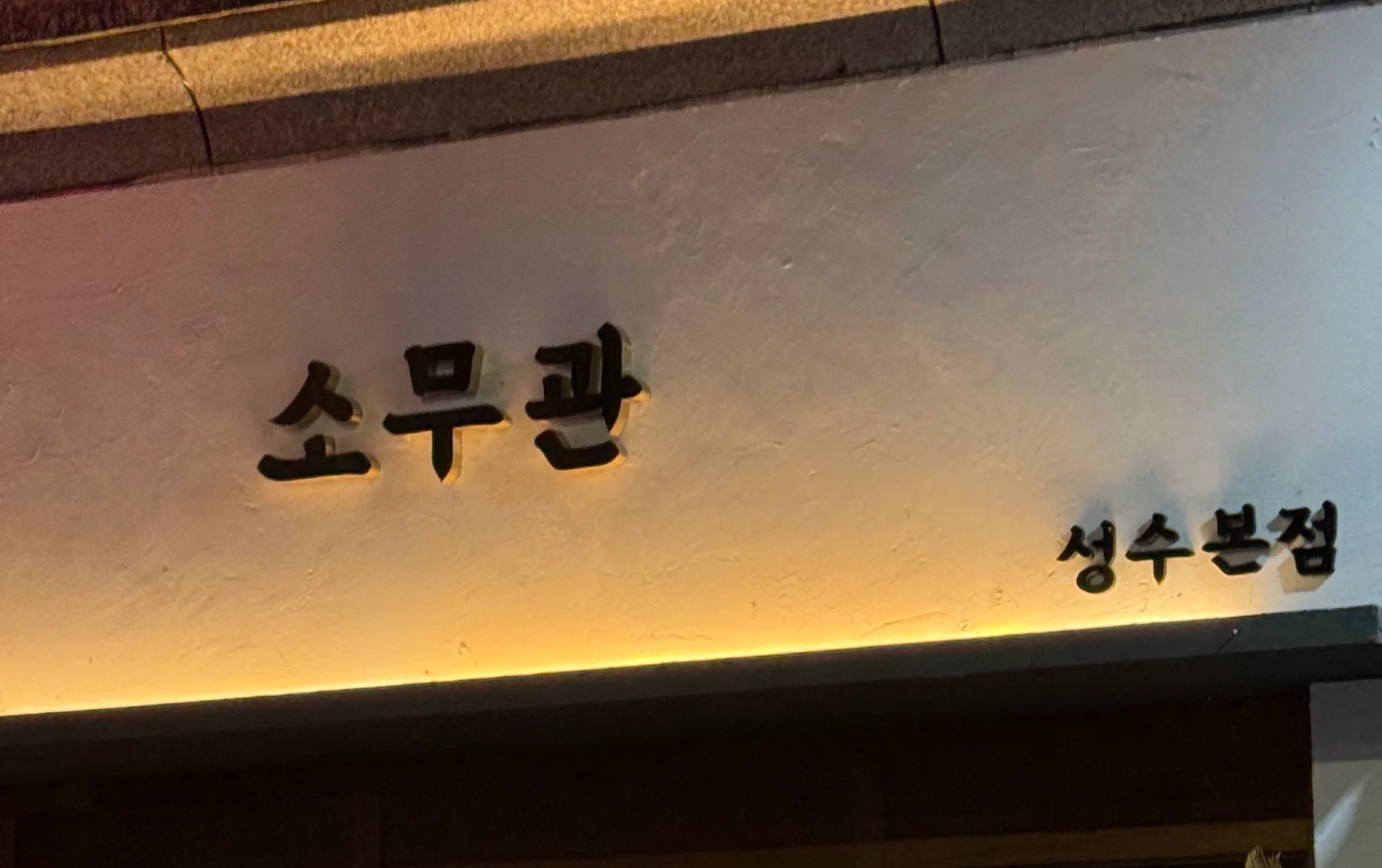 밤에 촬영한 소무관 성수본점 간판. 오프화이트 벽에 붙인 검은 각인 글자 「소무관」과 「성수본점」을 아래에서 앰버색 조명이 비춘다.