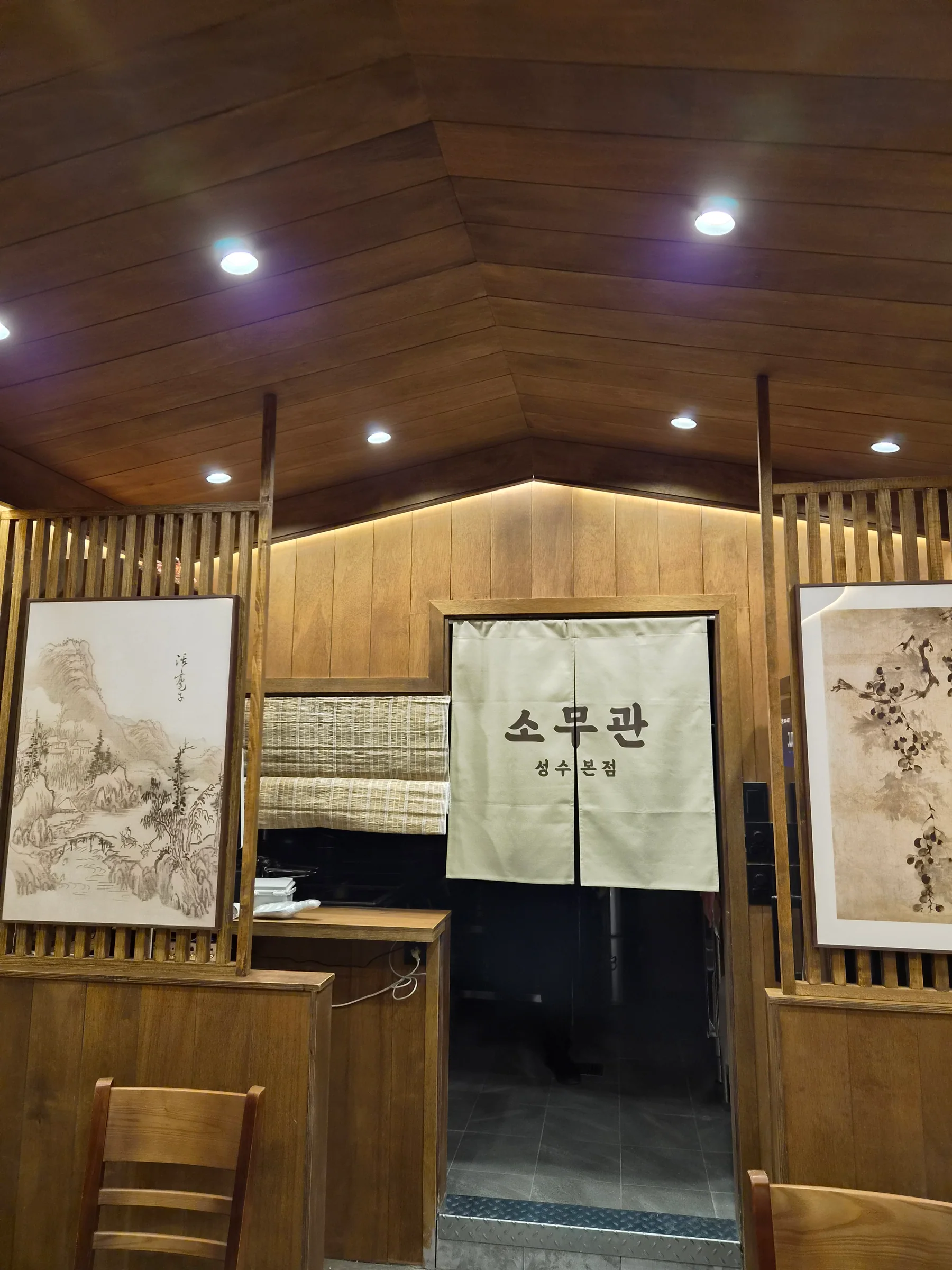 소무관 성수본점 내부. 꺾인 원목 천장 아래 세로 살 파티션이 서 있고, 주방 입구에 「소무관 성수본점」이라 쓰인 노렌이 걸려 있으며 양옆 벽에 수묵화 액자가 걸려 있다.